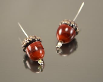 May include: A pair of acorn-shaped earrings with a red and brown glass bead and a silver cap. The earrings are on a white background.