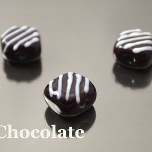 May include: Three chocolate candies with white stripes on top. The candies are arranged in a triangular pattern on a silver surface.