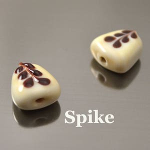 May include: Two white ceramic beads with brown leaf designs. The beads are shaped like a teardrop and have a hole in the top for stringing. The word "Spike" is written below the beads.