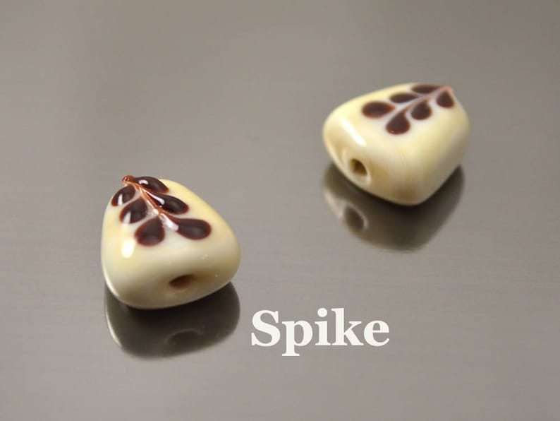 May include: Two white ceramic beads with brown leaf designs. The beads are shaped like a teardrop and have a hole in the top for stringing. The word "Spike" is written below the beads.