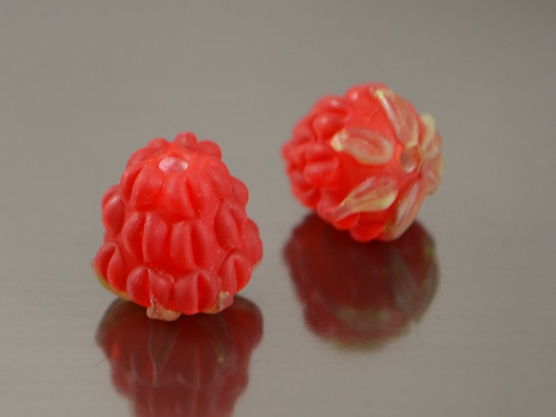 May include: Two red glass beads shaped like raspberries. The beads have a textured surface and a small hole in the center.