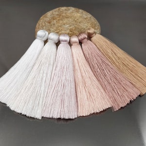 May include: Six silk tassels in various shades of pink and white. The tassels are arranged in a semi-circle on a brown stone surface.