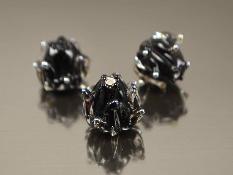 May include: Three small, black frog figurines with silver accents are arranged on a reflective surface. The frogs have detailed features, including textured skin and prominent eyes. The image is well-lit, highlighting the glossy finish of the frogs.
