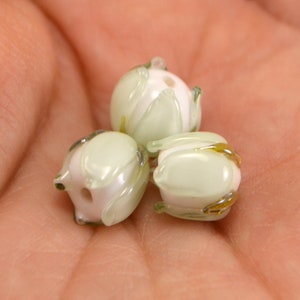 May include: Three glass beads in the shape of flowers. The beads are light green with white centers and have a translucent finish. The beads have a delicate, floral design.