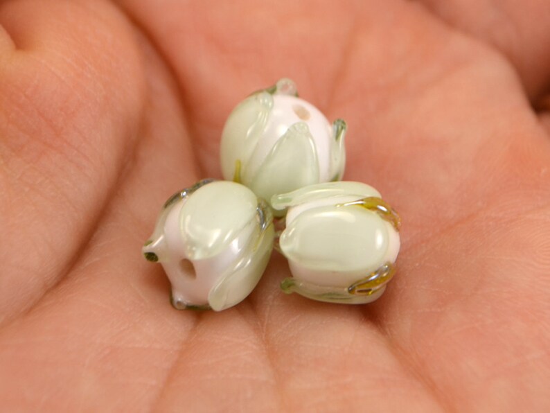 May include: Three glass beads in the shape of flowers. The beads are light green with white centers and have a translucent finish. The beads have a delicate, floral design.
