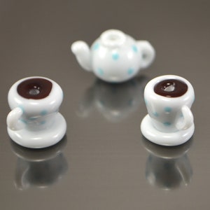 Puede incluir: Un juego de tres tazas de t&eacute; en miniatura y una tetera, todas blancas con lunares azules. Las tazas de t&eacute; contienen l&iacute;quido marr&oacute;n.