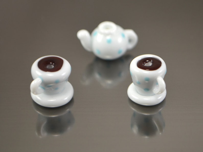 Puede incluir: Un juego de tres tazas de t&eacute; en miniatura y una tetera, todas blancas con lunares azules. Las tazas de t&eacute; contienen l&iacute;quido marr&oacute;n.