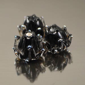May include: Three black glass frog figurines with silver and blue accents. The frogs have a glossy finish and are arranged in a cluster, with their reflections visible below. The overall theme is decorative art.