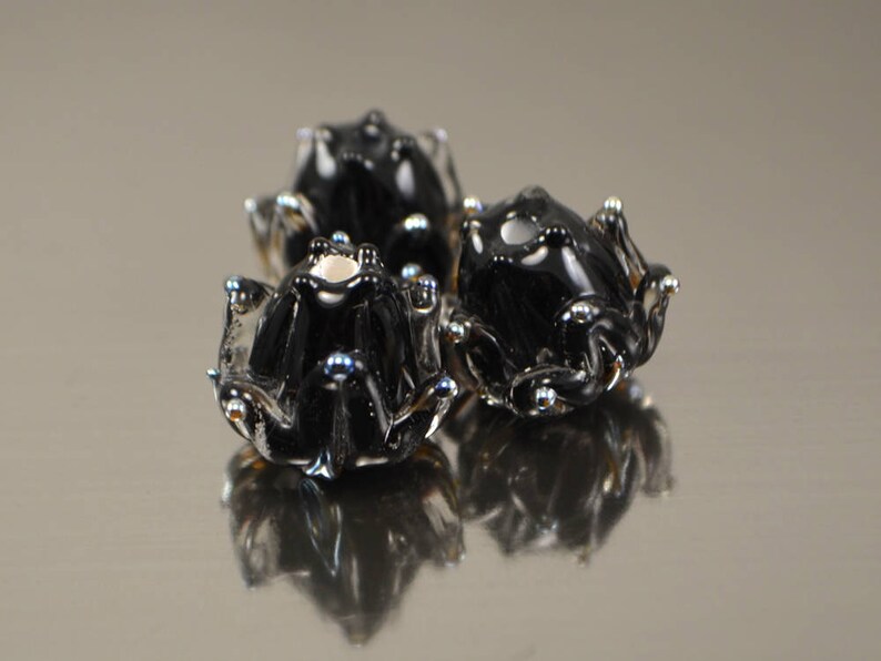May include: Three black glass frog figurines with silver and blue accents. The frogs have a glossy finish and are arranged in a cluster, with their reflections visible below. The overall theme is decorative art.
