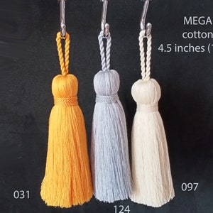 May include: Three cotton tassels hanging from hooks. The tassels are yellow, gray, and white. The tassels are 4.5 inches (12 cm) long. The text "MEGA cotton 4.5 inches (12 cm)" is at the top of the image. The numbers "031", "124", and "097" are below the tassels.