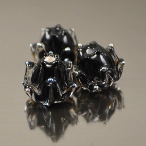 May include: Three black glass frog figurines with silver and blue accents. The frogs have a glossy finish and are arranged in a cluster, with their reflections visible below. The overall theme is decorative art.