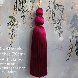 Puede incluir: Una borla decorativa de color magenta intenso con dos cuentas esféricas apiladas, una unión dorada y un lazo para colgar dorado. La borla mide 20 cm de largo y tiene un fleco grueso y denso. El texto dice: DECOR tassels 7.9 inches (20cm) MEGA thickness with beads golden binding and loop 150+ colors.