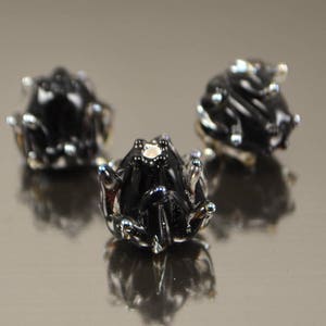May include: Three small, black frog figurines with silver accents are arranged on a reflective surface. The frogs have detailed features, including textured skin and prominent eyes. The image is well-lit, highlighting the glossy finish of the frogs.