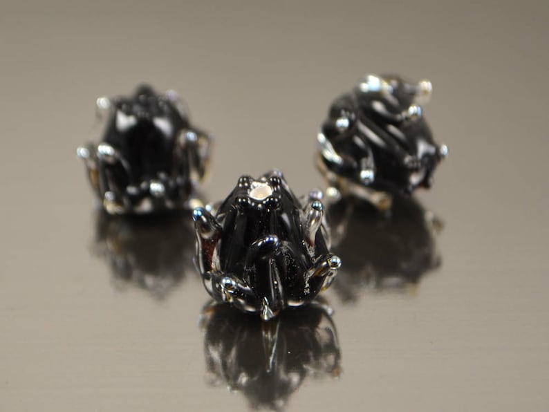 May include: Three small, black frog figurines with silver accents are arranged on a reflective surface. The frogs have detailed features, including textured skin and prominent eyes. The image is well-lit, highlighting the glossy finish of the frogs.