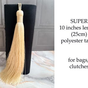 May include: A beige polyester tassel, 10 inches long, hangs in front of a black object. The tassel has a woven top and long, flowing strands. Text on the right reads "SUPER 10 inches length (25cm) polyester tassel for bags, clutches."