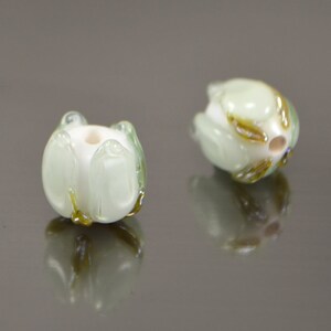 May include: Two white glass beads with green and gold accents, shaped like flowers. The beads are on a reflective surface.