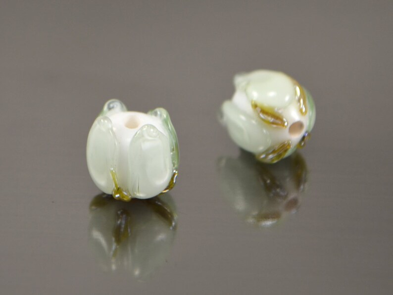 May include: Two white glass beads with green and gold accents, shaped like flowers. The beads are on a reflective surface.