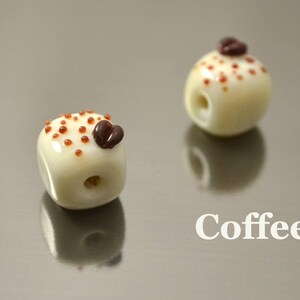 May include: Two white ceramic beads with brown coffee bean accents. The beads are decorated with brown dots. The beads are on a silver surface. Coffee.