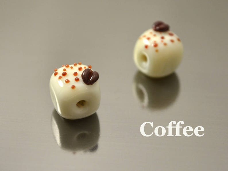 May include: Two white ceramic beads with brown coffee bean accents. The beads are decorated with brown dots. The beads are on a silver surface. Coffee.