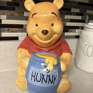 Puede incluir: Un tarro de cerámica para galletas con forma de oso amarillo que lleva una camiseta roja. El oso sostiene un tarro de miel azul con la palabra HUNNY impresa en letras negras en el frente, con un pequeño detalle de abeja y miel amarilla.