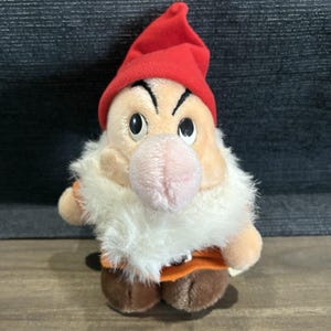 May include: A plush toy of Grumpy from Snow White. The toy has a red hat, a white beard, and an orange shirt. The dwarf has a pink nose and black eyebrows. The toy is standing on a wooden surface.