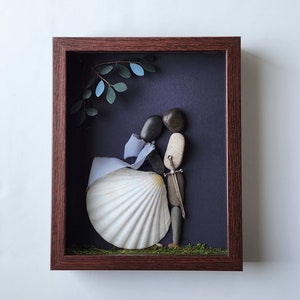 May include: A framed artwork featuring a couple made of stones, a seashell, and a small sword. The couple is standing on a bed of moss, with a blue and green leafy branch above them. The background is a dark purple.