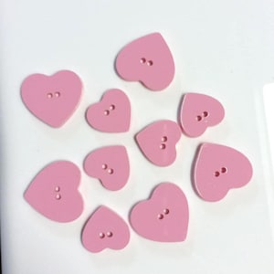May include: A selection of light pink heart-shaped buttons. Each button features two or three holes for sewing. The buttons are arranged on a white surface. These buttons are suitable for sewing, crafting, and adding a touch of charm to various projects.