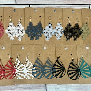 May include: Assortment of dangle earrings with geometric designs. The earrings are displayed on brown card stock. The earrings are in various colors, including red, white, black, gray, and green.