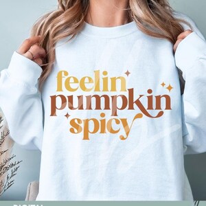 May include: A light blue sweatshirt with the text "feelin' pumpkin spicy" in orange, brown, and yellow lettering. The text is in a stylized font with a glittery effect. The sweatshirt is a digital download available in SVG, PNG, and DXF formats.