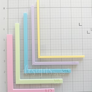 May include: Five L-shaped plastic guides in pastel colors including pink, light green, blue, lavender, and yellow. Each guide features an embossed fraction measurement: 1/2, 3/8, 5/16, and 1/4 inches, arranged on a grid mat for scale and alignment.