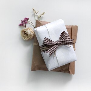 May include: A white gift box wrapped in brown paper with a brown and white gingham ribbon. The box is decorated with dried flowers.