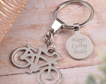 May include: A silver-colored keychain featuring a bicycle silhouette and a round tag. The tag is engraved with the words "Happy Cycling Buddy". The keychain is on a wooden surface.