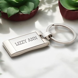 May include: A silver rectangular keychain with the engraved text "LIZZY ANN". The keychain has a rounded edge and a circular key ring. The background includes small potted plants.