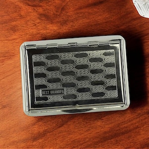 May include: A silver metal cigarette case with a decorative woven pattern and the words "BEST GRANDPA" engraved on the front. The case is rectangular with rounded corners and a hinged lid. The case is on a wooden surface.