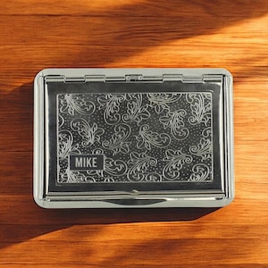 May include: A silver business card holder with an engraved floral design. The name "MIKE" is displayed in a small box on the front. The rectangular holder has rounded corners. The item is on a wooden surface.