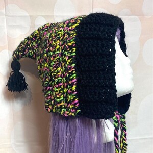 May include: A handmade crochet hat with a pointed tip and a black tassel. The hat features a black hood and a colourful pattern of neon yellow, pink, and green yarn. The hat is designed to be worn.