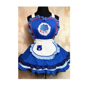 May include: Blue apron with a heart-shaped bodice featuring a cloud and rain design. The apron has a white bib with a bear graphic and is trimmed with ruffles. The straps are white, and the overall design is reminiscent of a cartoon character.