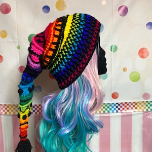 May include: A colorful, handmade crochet hat with a long, pointed tail. The hat features a rainbow gradient of colors, including red, orange, yellow, green, blue, and purple. The tail is adorned with small, colorful bobbles and a black tassel. The hat is a whimsical accessory.
