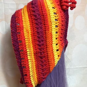 May include: A handmade crochet hood in vibrant colors. The hood features a gradient of red, orange, and yellow stripes. The hood has a textured stitch pattern and a decorative spiral at the top. The hood is shown on a purple wig.