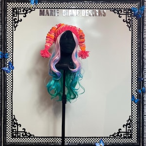 May include: A vibrant crochet hat in shades of pink, orange, and white, with two pom-poms. The hat is displayed on a black mannequin head with a wig of pink and teal hair. The background includes the text "MARIE DIANE DESIGNS".