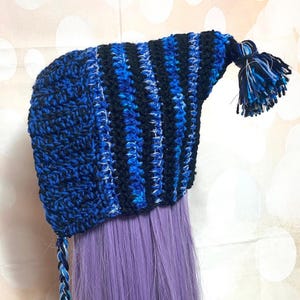 May include: A handmade crochet hat in shades of blue and black. The hat features a striped pattern on the top and a solid blue texture on the front. A tassel hangs from the back, and a braided tie is on the side.