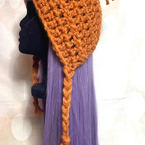 May include: An orange crochet hat with a pointed top and braided tassels. The hat is displayed on a black mannequin head with purple hair in the background. The hat's texture is visible.