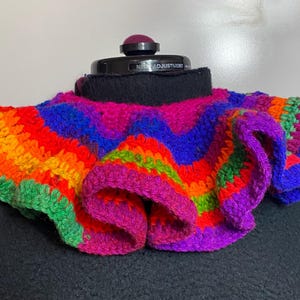 May include: A colorful, crocheted collar in a rainbow of hues, including green, orange, red, purple, and blue. The collar is ruffled and appears to be handmade. It is displayed on a black mannequin.