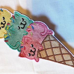 May include: An embroidered patch of three ice cream scoops shaped like cats in a waffle cone. The scoops are yellow, green, and pink with sprinkles.