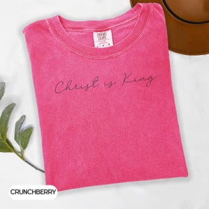 A pink t-shirt with the text "Christ is King" printed on it, along with a brown hat and some greenery.