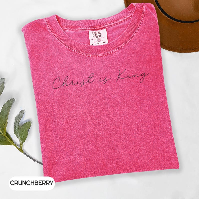 A pink t-shirt with the text "Christ is King" printed on it, along with a brown hat and some greenery.