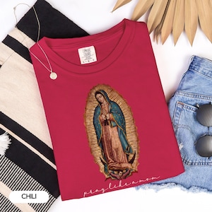 May include: A red t-shirt featuring an image of Our Lady of Guadalupe, with a blue cloak and gold halo. The text "pray like a mom" is below the image. The shirt is styled with denim shorts and a gold necklace.
