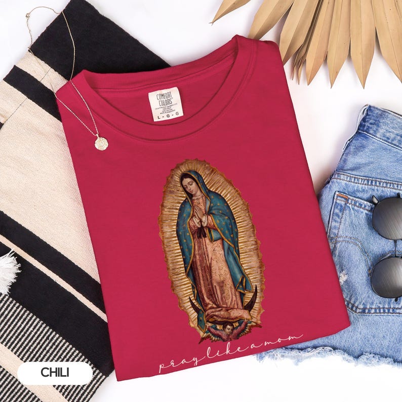 May include: A red t-shirt featuring an image of Our Lady of Guadalupe, with a blue cloak and gold halo. The text "pray like a mom" is below the image. The shirt is styled with denim shorts and a gold necklace.
