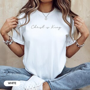 A woman wearing a white t-shirt with the text "Christ is King" printed on it, posing with her hands behind her back.