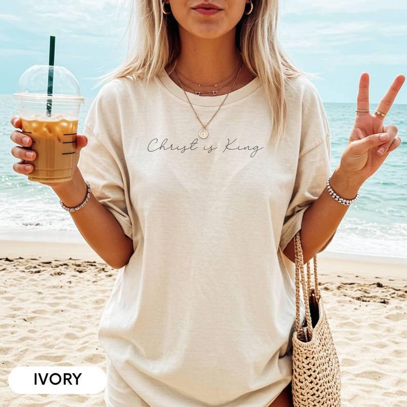 A woman wearing a white t-shirt with the text "Christ is King" printed on it, standing on a beach with her hands in a peace sign and holding a drink.